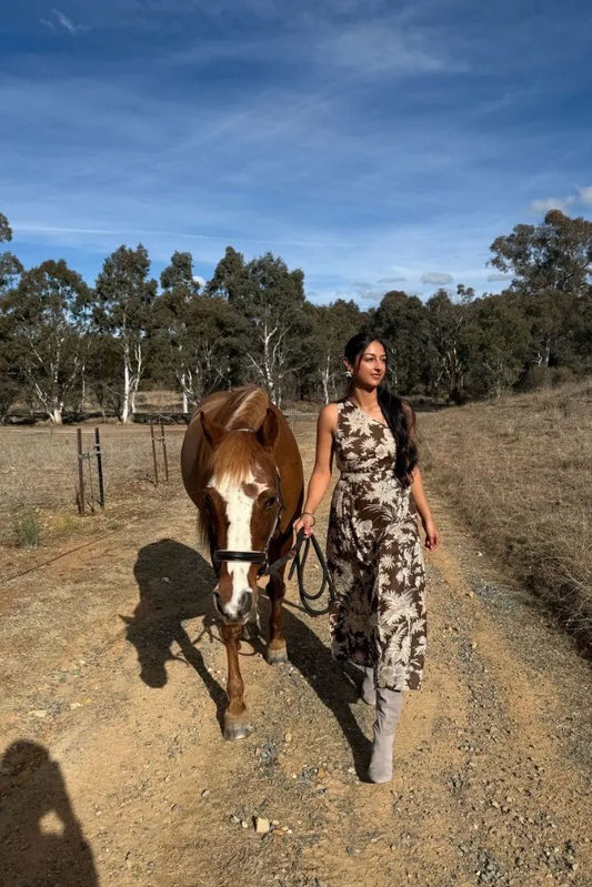 Harnoop in Cocoa One Shoulder Midi walking a horse on a dirt path with trees and blue sky in the background