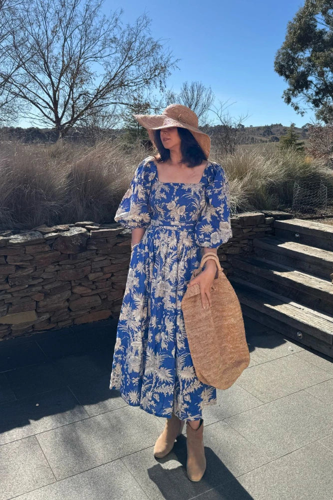 Tina wearing the blue Eliza Midi dress with a straw hat and bag standing outdoors.